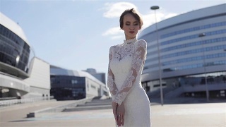 美丽的模特在优雅的婚纱站在商业城市中心街道建筑的背景。年轻微笑的女子在优雅的白色蕾丝设计长袍享受阳光在城市周围视频素材模板下载