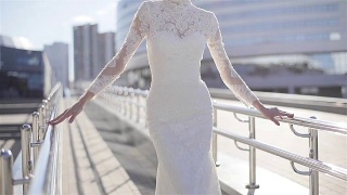 女人在优雅的白色长蕾丝设计连衣裙摆姿势在阳光明媚的城市背景春天快乐。新娘在城市街道上近距离展示昂贵的服装。婚纱时装设计师服装视频素材模板下载