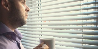 年轻的商人站在窗边喝咖啡。在办公室。一个严肃的人坐在窗外喝茶。深思熟虑的问题。老板喝咖啡，喝茶。疲倦、悲伤、沮丧的人会休息。