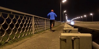 晚上在桥边慢跑的运动员。一个穿着运动服的男人在晚上跑步。男正在健身，车路过。傍晚沿着高速公路慢跑。