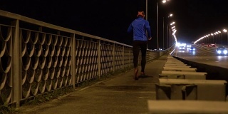 晚上在桥边慢跑的运动员。一个穿着运动服的男人在晚上跑步。男正在健身，车路过。傍晚沿着高速公路慢跑。
