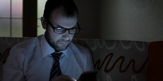 一个穿着白衬衫、打着蓝领领带的严肃男人，晚上在家用智能手机工作。商人使用智能手机，晚上坐在沙发上。