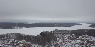 冬村鸟瞰图，河岸边有输电铁塔。冬季景观上的无人机输电塔和电力线