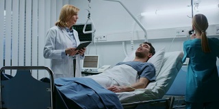 友好的女医生访问微笑的康复男子躺在床上，她问他问题和填写医疗图表，护士检查他的生命体征。