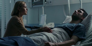 在医院里，忧心忡忡的妻子坐在她生病的丈夫的床边。她握着他的手，和他说话，希望他能痊愈。