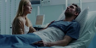 在医院里，一个病人躺在病床上，他来访的妻子满怀希望地坐在他旁边，为他的早日康复祈祷。悲惨、忧郁和忧郁的场景。
