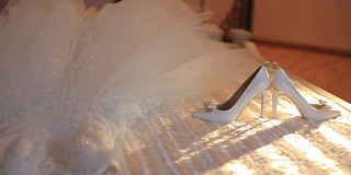 近距离观看结婚戒指之间的新娘高跟鞋躺在床上的裙子。移动的阳光。