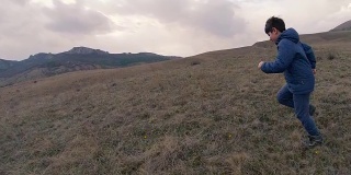 一个男孩在长着高草的场地上跑向群山环绕的地平线。慢动作视频