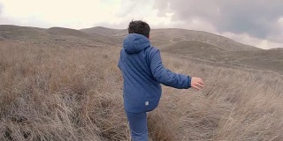 一个在长着高草的场地上的男孩跑到有山的地平线上。慢动作视频