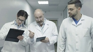 三名男医生走在医院走廊上讨论病人的笔记。FullHD视频素材模板下载