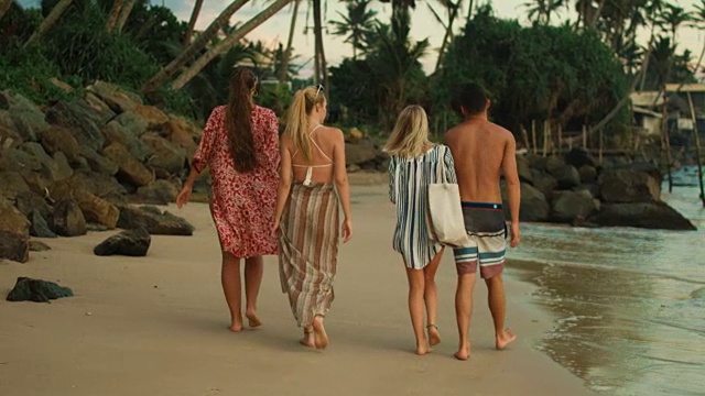三个漂亮的女孩和一个英俊的男人在海滩上散步。甘美的绿色森林与棕榈树在他们之前。