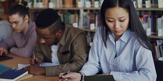 集中的亚洲女孩学生准备考试和写笔记，而坐在桌子上的大学图书馆与她的同学学习书籍