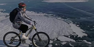 一个女人在冰上骑自行车。这个女孩穿着银色的羽绒服，背着自行车背包，戴着头盔。用四旋翼无人机拍摄。自行车的轮胎上布满了钉子。