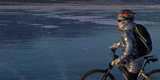一个女人在冰上骑自行车。这个女孩穿着银色的羽绒服，背着背包，戴着头盔。骑自行车的人一边骑一边停下来休息。他坐在方向盘上，取下部分齿轮。