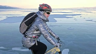 一个女人在冰上骑自行车。这个女孩穿着银色的羽绒服，背着自行车背包，戴着头盔。结冰的贝加尔湖的冰。自行车的轮胎上覆盖着特殊的尖钉。视频素材模板下载