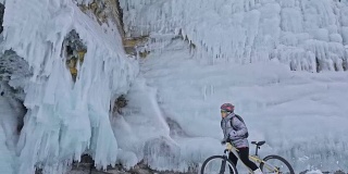 一个女人在冰洞附近骑自行车。有冰洞和冰柱的岩石非常漂亮。这个女孩穿着银色的羽绒服，背着自行车背包，戴着头盔。旅行者正在骑自行车。
