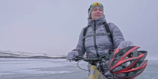 女人穿着运动器材。这个女孩穿着银色的羽绒服，背着自行车背包，戴着头盔。结冰的贝加尔湖的冰。自行车的轮胎上覆盖着特殊的尖钉。