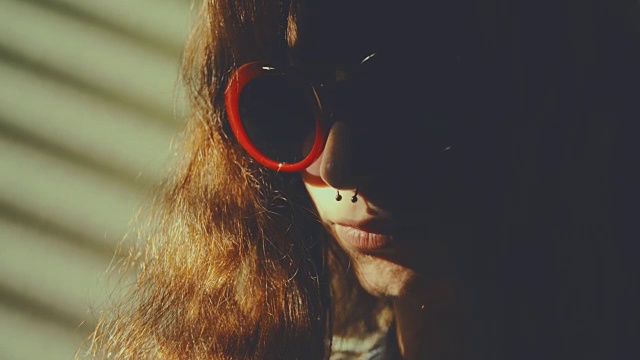 透过百叶窗的光线，映入眼帘的是一个戴着红色太阳镜的穿孔女孩，她正在用智能手机冲浪。慢动作拍摄