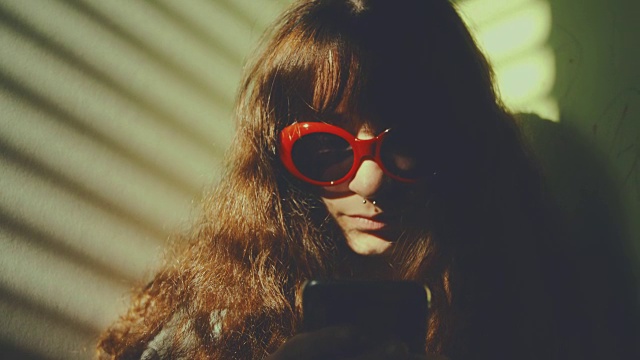 透过百叶窗的光线，映入眼帘的是一个戴着红色太阳镜的穿孔女孩，她正在用智能手机冲浪。慢动作拍摄