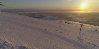 在阳光明媚的冬季日落，人们乘坐缆车在滑雪坡上滑雪和滑板。太阳在坐标系。鸟瞰图。无人机正在横向飞行。远景。