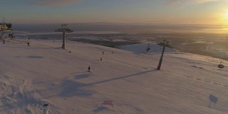 在阳光明媚的冬季日落，人们乘坐缆车在滑雪坡上滑雪和滑板。鸟瞰图。无人机正在横向飞行。远景。