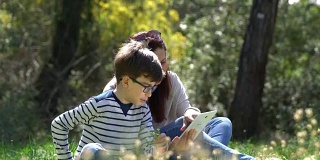 美丽的女人和她的儿子坐在森林里，使用数字平板电脑