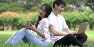 情侣们拿着智能手机和笔记本电脑躺在户外公园的草地上。男朋友和女朋友在假期快乐和微笑放松。