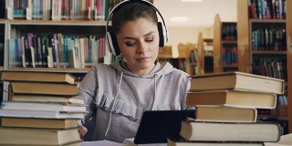 一位戴着白色耳机、面带微笑的年轻女子正坐在大学图书馆的桌子旁，手里拿着平板电脑，听着音乐。她在跳舞，在笔记本上写讲座