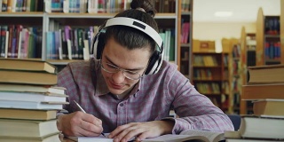 年轻的白人男学生戴着大耳机坐在图书馆的桌子旁。他周围有很多书，他在微笑着听音乐和写作