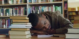 非裔美籍英俊年轻男学生正坐在图书馆里堆满书的桌子旁。他的头躺在桌子上，他正在睡觉，很累。