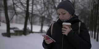漂亮的黑发女人在雪地上喝咖啡，用手机，冬天