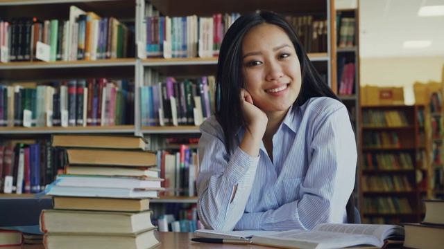 年轻美丽的亚洲女学生的肖像坐在桌子与图书馆的教科书看着相机。她满脸堆笑。