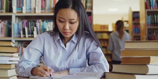 年轻美丽的亚洲女学生坐在桌子周围的书籍在大图书馆重写课本准备考试