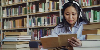 相当积极的亚洲青少年学生戴着耳机听音乐坐在大图书馆的桌子上拿着书和写总结