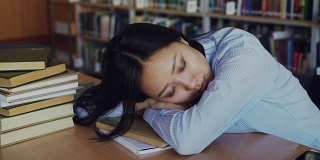 年轻美丽疲惫的亚洲女学生坐在书桌上，她的头在它上，她是睡在教科书周围的堆在大大学图书馆