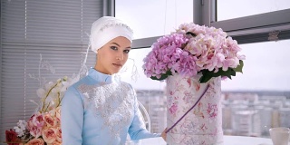 美丽的穆斯林新娘与蓝色的传统服装尼卡坐在窗户附近