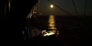 一名女子在满月背景下的帆船上使用4K智能手机