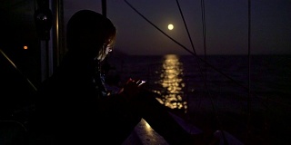 在宁静的满月照耀海洋的夜晚，4K女人在帆船上使用智能手机，实时