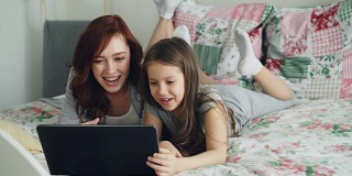 快乐可爱的女儿和年轻的母亲躺在舒适的卧室床上用笔记本电脑与父亲或祖母进行在线视频通话
