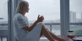 年轻女子享受着她的早茶，看着窗外的雨。美丽浪漫的陌生女孩在舒适的家里喝热饮。雨天的心情