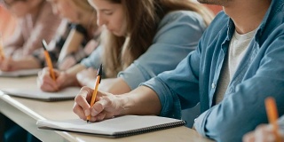 一排多民族学生在教室里考试的移动镜头。专注于拿笔和在笔记本上写作。聪明的年轻人在大学学习。