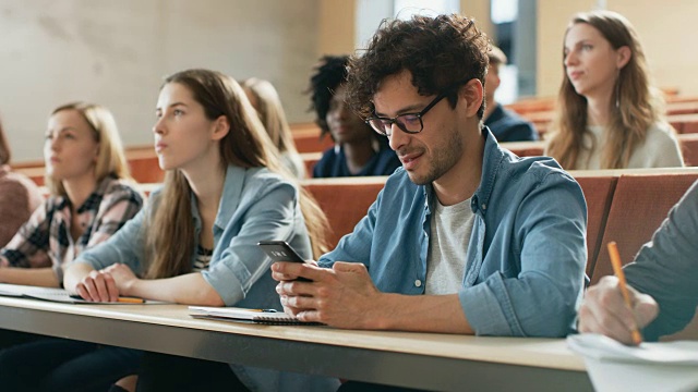 西班牙裔男子在课堂上讲课时使用智能手机。报告厅里坐满了正在学习的学生。大学里的年轻人。
