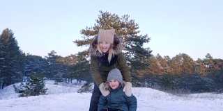 冬日里，快乐的年轻女子推着雪橇把儿子从雪山上拉下来，慢慢地跟在他后面跑
