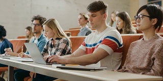 英俊的白人学生在大学听讲座时使用笔记本电脑。多元民族的现代聪明学生投资他们的未来。