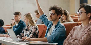 英俊的西班牙裔学生在大学听讲座时使用笔记本电脑，他举手问讲师一个问题。现代聪明学生的多民族群体。