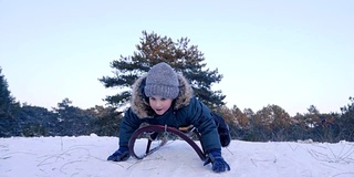 快乐的男孩与举起的手躺在雪橇上，骑从雪山在冬天的森林在缓慢的动作