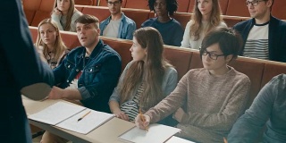 镜头面对课堂:大学教授给满教室的多民族学生讲课的背影。当学生听时，讲师用手打手势。