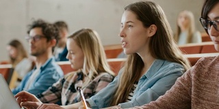 年轻漂亮的学生在大学听讲座时使用笔记本电脑，她举手向讲师提问。现代聪明学生的多民族群体。