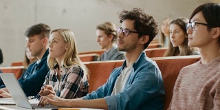 英俊的西班牙裔学生在大学听讲座时使用笔记本电脑，他举手问讲师一个问题。现代聪明学生的多民族群体。