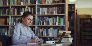 美丽严肃的白人女学生坐在桌子上的书在图书馆用笔记本电脑在抄写总结